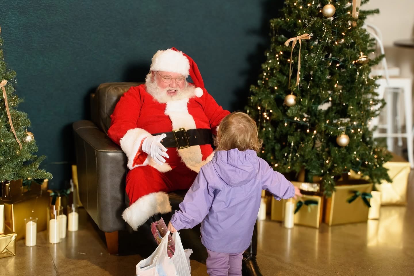 Santa Michael laughing in a leather armchair as a toddler in a purple jacket runs to greet him beside a Christmas tree