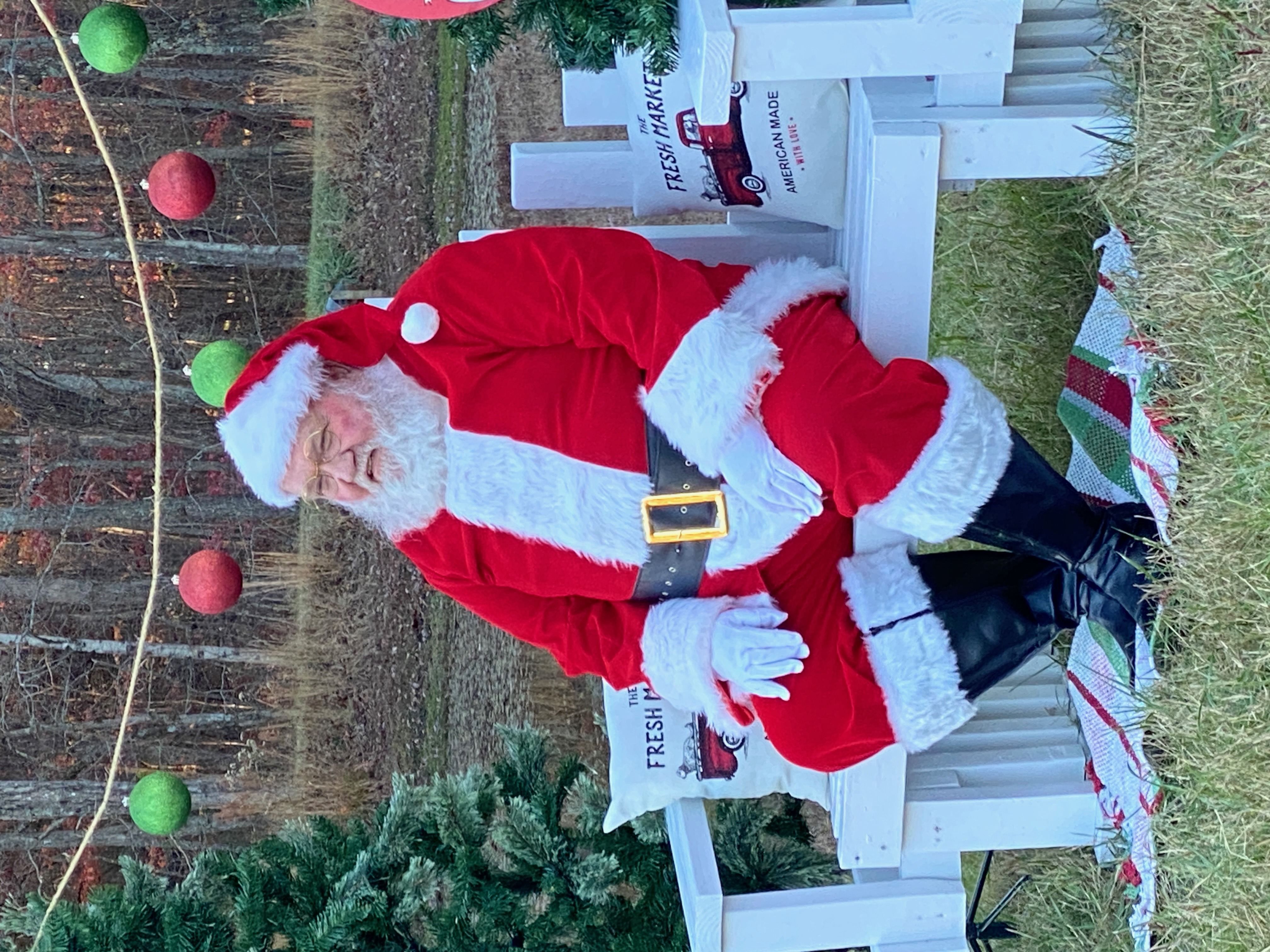 Santa Michael seated on a white bench outdoors under strung red and green ornaments, autumn trees behind him